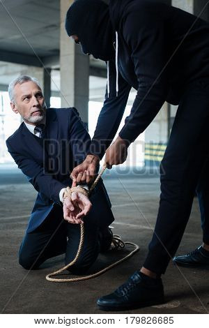 Kidnapping victim. Cheerless scared elderly man sitting on the floor and looking at his kidnapper while having his hands tied