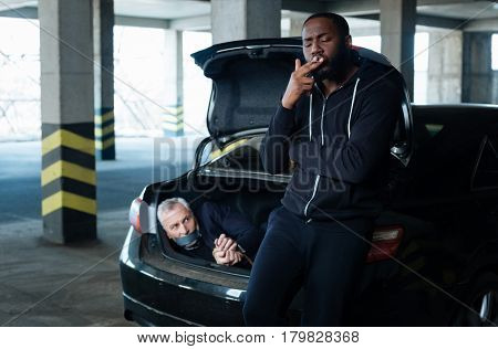 Unhealthy habit. Nice relaxed adult criminal leaning on the car boot and smoking a cigarette while having a tied man in it