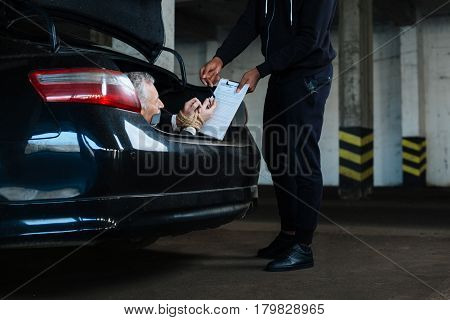 Frightened to death. Cheerless frightened aged man taking a pen and signing a document while being kidnapped