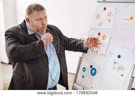 A little bit sick. Attentive unhealthy stout man pinning some diagrams on a board and working on his business presentation while coughing