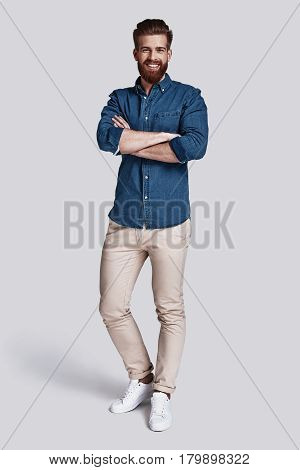 Confident in his style. Full length of handsome young man looking at camera and keeping arms crossed while standing against grey background