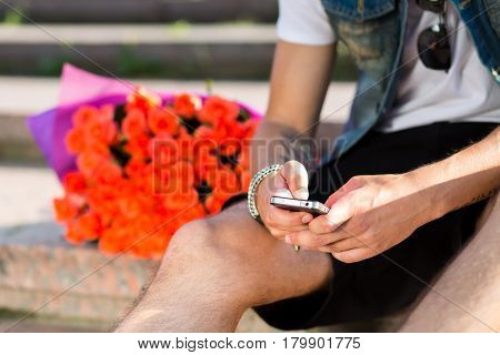 Waiting for you. Young man typing message on the mobile phone while sitting on the staircase with bouquet of roses laying near him