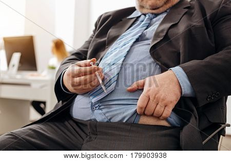 Needed remedy. Scrupulous pudgy unhealthy man doing an injection at his workplace because of not eating healthy and having excess weight