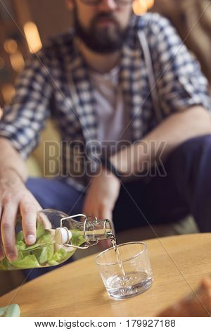 Young hipster guy sitting in an armchair in a cafe pouring a glass of detox water with mint leaves