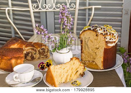 Typical italian flavorous home baked cakes for Easter: La Gubana with sophisticated stuffing and Panettone with candied flowers on the table covered for tea time. Horizontal. Main colors: white gray yellow brown.