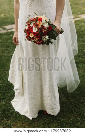 Beautiful bride holding wedding bouquet stylish bride in lace white dress holding red roses bouquet in her hand closeup posing outdoors near church