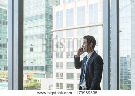 Mature and confident business executive looking looking out of large windows at a view of the city below from the top floor of an office building while talking on his mobile phone