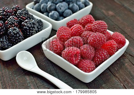 Mix of blueberries, blackberries and raspberries fruit on wood table. Forest fruit in white idaeus with spoon. Delicious fresh berries.