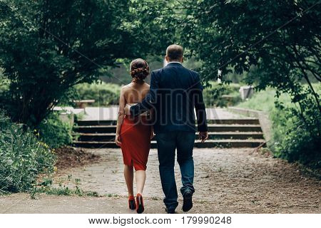 Romantic Newlywed Couple Walking In Park, Handsome Goom Holding Hand Around Waist Of Sexy Bride In R