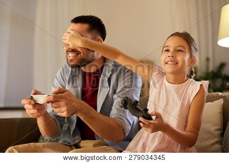 family, gaming and entertainment concept - happy father and little daughter with gamepads playing video game at home in evening