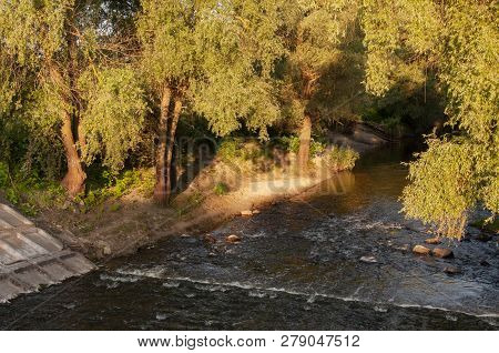 Small River Slow Flowing Across The Green Forest With Reflections Of  Trees In The Water.  Summer