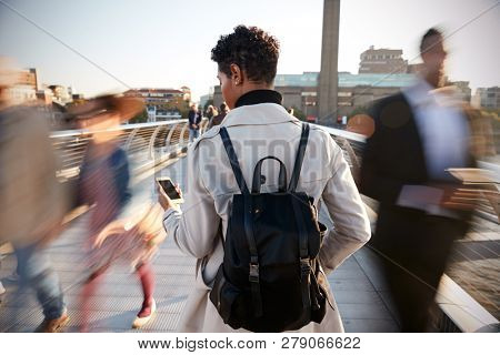 Back view of young adult woman standing on Millennium Bridge, London, using smartphone, motion blur