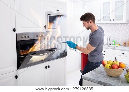 Man Using Fire Extinguisher To Stop Fire Coming Out From Oven In Kitchen