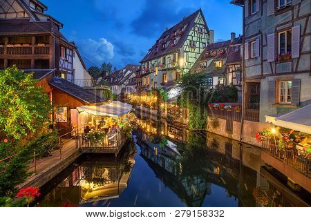 Colmar, France. Half-timbered Houses And Verandas Of Restaurants Reflecting In The Water At Dusk In 