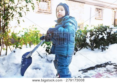 Little Caucasian Boy Shovels Snow In The Yard With Beautiful Snowy Rose Bushes. Child With Shovel Pl