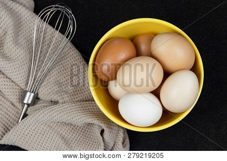 Top View Of Hen Eggs In A Yellow Bowl And An Eggbeater On A Black Background.