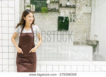 Portrait Of A Happy Young Asian Barista In Apron Smiling And Looking Away In Front Of Her Small Coff