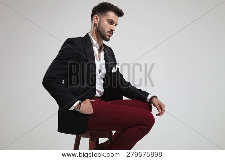 side view of seated smart casual man wearing black suit and red pants looking down on light grey background, portrait picture