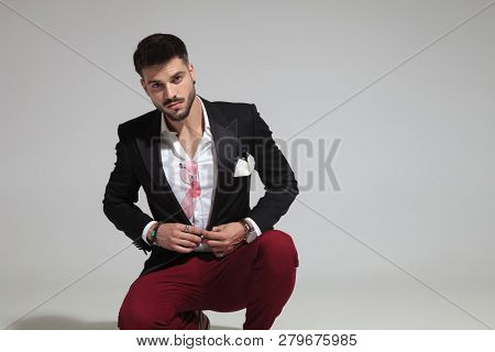 sexy smart casual man crouching on light grey background and buttoning black suit, portrait picture