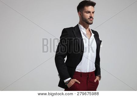 portrait of handsome smart casual man holding pockets and looking to side while standing on light grey background