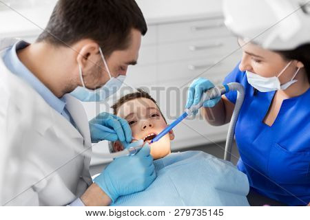 medicine, dentistry and healthcare concept - dentist and assistant with dental drill and saliva ejector treating kid patient teeth at dental clinic