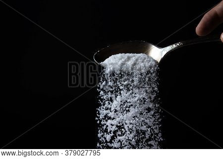 Salt Is Poured From A Spoon On A Black Background. Excessive Salt Intake. Coarse Salt
