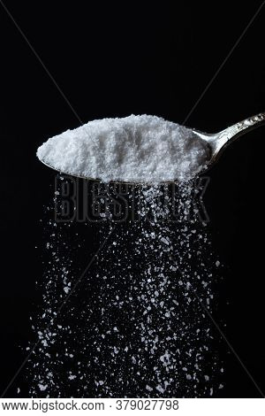 Salt Is Poured From A Spoon On A Black Background. Excessive Salt Intake. Coarse Salt