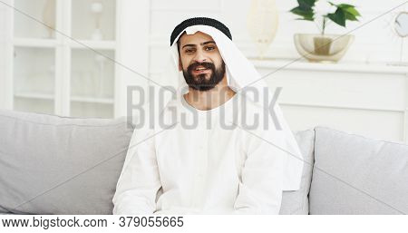 Portrait Of Handsome Young Arabian Man In Kandura Sitting On Couch In Living Room And Smiling To Cam