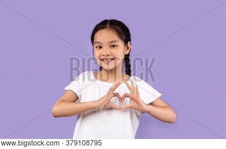 Love And Kindness. Little Asian Girl Gesturing Heart Shape Smiling To Camera Posing On Purple Studio
