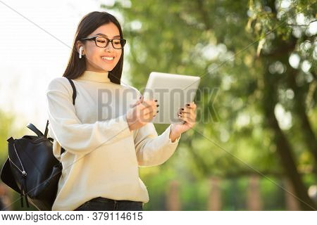 Technology And Communication Concept. Happy Asian Girl Wearing Specs And Earbuds Holding Tablet, Out
