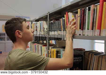 Italy, Summer 2020: A Caucasian Boy Is Consulting Books In A Public Library. High Quality Photo