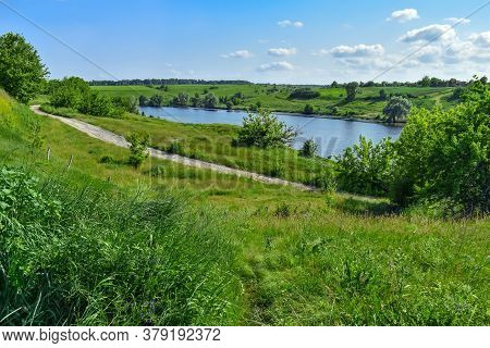 Dirt Road Among Green Hills On The Shore Of A Pond With Blue Water. Bright Natural Panorama With A L