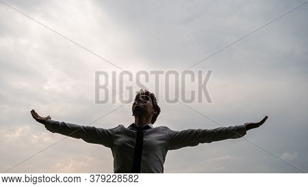 Low Angle View Of A Young Businessman Standing Under Cloudy Grey Sky Aith His Arms Spread Widely.