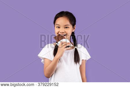 Sweet Tooth. Little Asian Girl Eating Chocolate Smiling To Camera Standing On Purple Background. Stu
