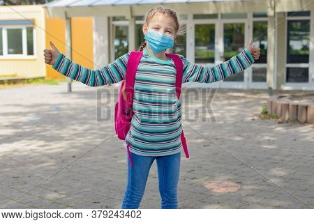 A Happy, Cute Caucasian Girl With A Pink Backpack Stands In Front Of The School Doors In A Protectiv