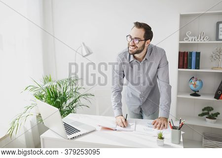 Smiling Young Bearded Business Man In Gray Shirt Standing Near Desk Working With Laptop Computer Pap
