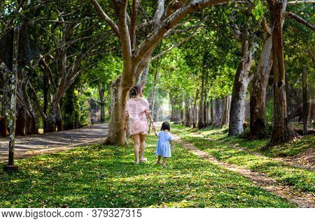 Mother Holding Hand Of Little Daughter And Walked Happily Around In The Garden Have Big Trees To Giv
