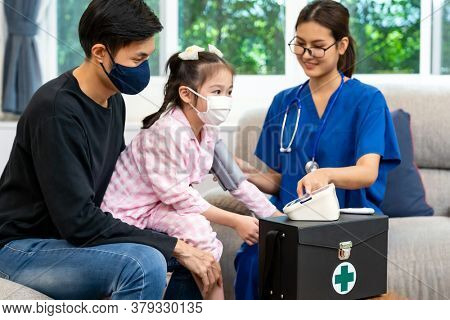 Asian doctor take blood pressure measurement to little girl child sit with her father in living room while doctor visit home. Home healthcare delivery concept.