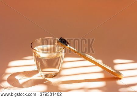 Bamboo Toothbrush And Glass Of Water On A Peach-colored Pastel Background With Shadows Of Natural Li