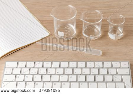 Glass Measuring Beakers Standing Gradually On Table And Plastic Pipette In Front Of A White Computer