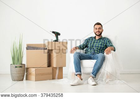 Portrait of smiling relaxed young bearded man sitting in armchair covered with napkin in new house with moving boxes