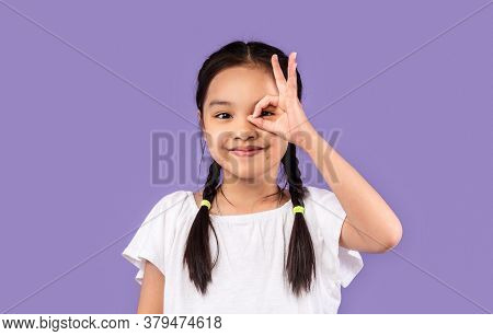Korean Little Girl Looking Through Okay Sign Covering Eye Posing On Purple Studio Background. Everyt