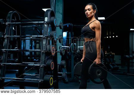 Side View Of Gorgeous Brunette Female Bodybuilder With Braids Posing Near Stand With Sports Equipmen