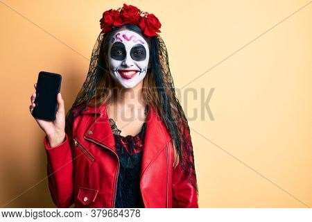 Woman wearing day of the dead costume holding smartphone showing screen looking positive and happy standing and smiling with a confident smile showing teeth 