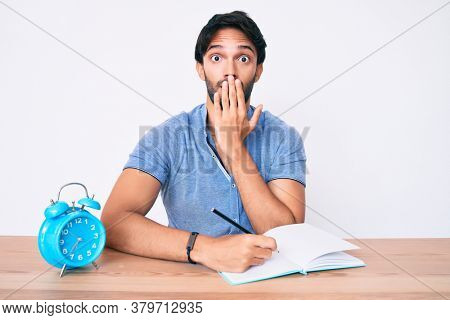 Handsome hispanic man sitting on the table stuying for university covering mouth with hand, shocked and afraid for mistake. surprised expression 