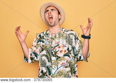 Young handsome man with blue eyes on vacation wearing summer florar shirt and hat crazy and mad shouting and yelling with aggressive expression and arms raised. Frustration concept.