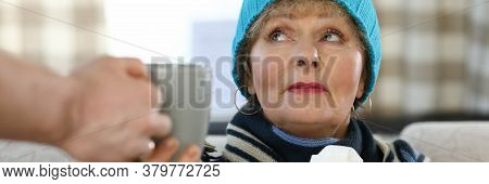 Elderly Woman In Scarf And Hat Sits On Sofa Home. Elderly Lady Takes Cup Tea. Weak Immune System In 