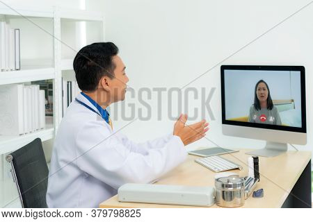 Asian Man Doctor Wears White Coat And Headset Speaking Videoconferencing On Laptop Computer Using On
