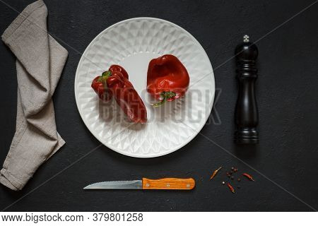 Two Ugly Red Peppers On A White Plate, A Pepper Mill, A Linen Napkin And A Knife, On A Black Backgro