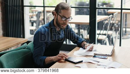Caucasian Young Man Sitting At Table And Doing Business Calculations. Man In Glasses Calculating Spe